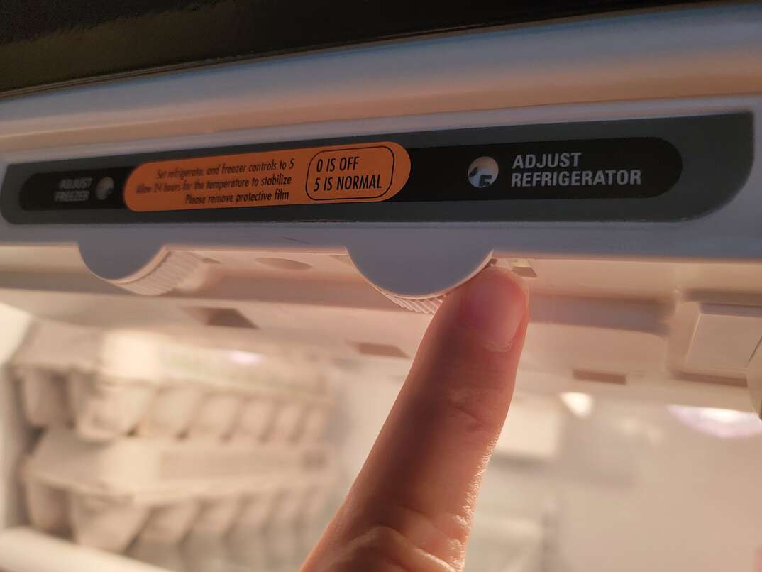 A person s finger adjusts the temperature control of a refrigerator, just inside the open fridge door, with two cartons of eggs visible in the background.