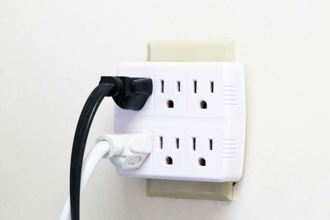Two plugs in North American electrical outlet with extension for six sockets on white wall. Electric multi splitter with several few cables plugged into it.