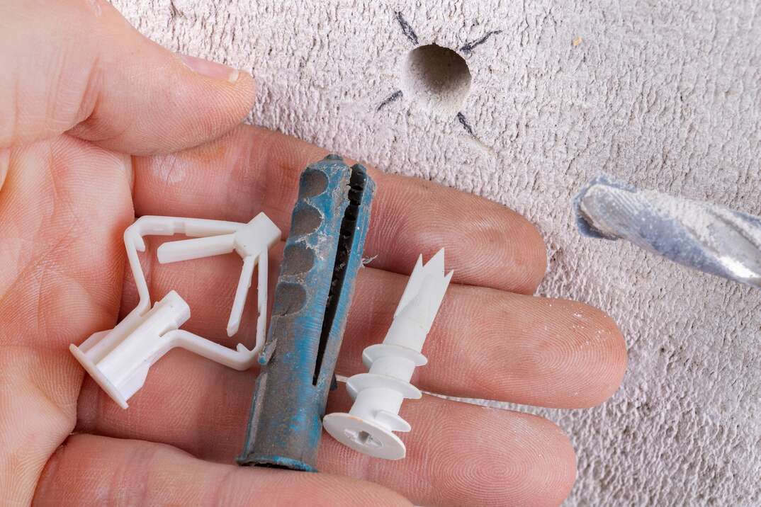 a hand holds 3 different types of wall anchors in front of a recently drilled hole inconcrete
