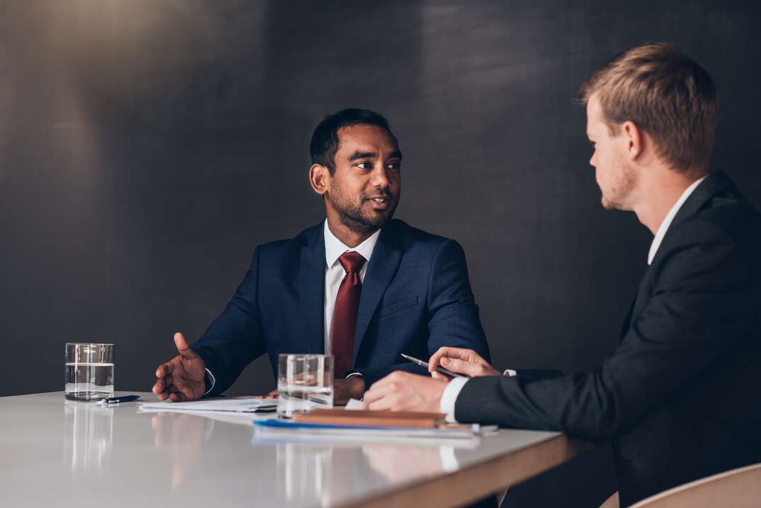 Two young businessmen in suits discussing documents together while sitting at a table in a modern boardroom