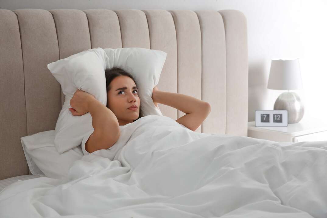 A woman lies in a bed with white sheets and a tall beige padded headboard holding white pillows over here ears to muffle noise as she tries to sleep, white sheets, bedding, white bedding, bed, pillow, headboard, white background, clean sheets, woman, lying in bed, in bed, sleeping, trying to sleep, holding pillows over ears