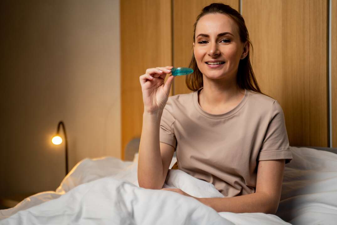 A young woman sits upright in bed with her legs under the covers up to her waist and is holding her dental night guard as a bedside lamp glows in the background, night guard, dental guard, dental night guard, lamp, nightstand, bedside lamp, bed, in bed, bedding, under the overs, woman, young woman, dental, dentist, teeth, oral health, dental health, medical device, dental device, heathcare, health care