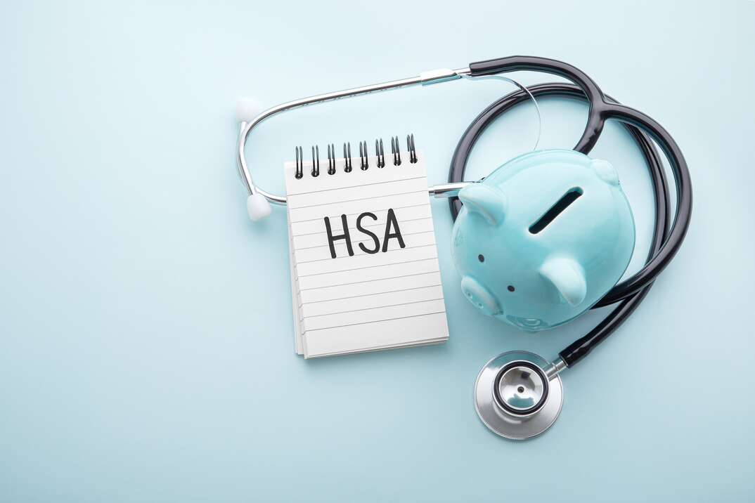 A light blue colored piggybank and a stethoscope and a notepad with the acronym HSA written on it sits against a light blue background, piggybank, piggy bank, light blue colored piggy bank, stethoscope, light blue background, notepad, notebook, note paper, HSA, health savings account, health insurance, health coverage, medical insurance, medical coverage, medical, health