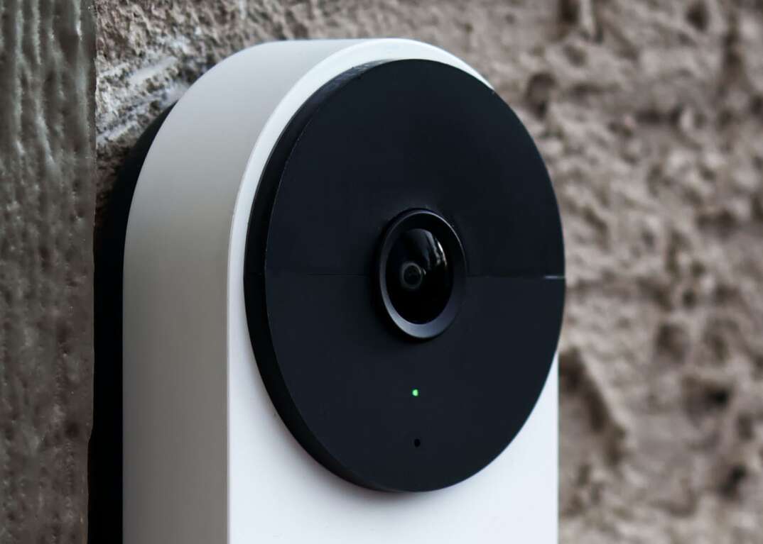 A white Google Nest Doorbell video security device with a black circular camera is affixed to a brown stucco exterior wall.