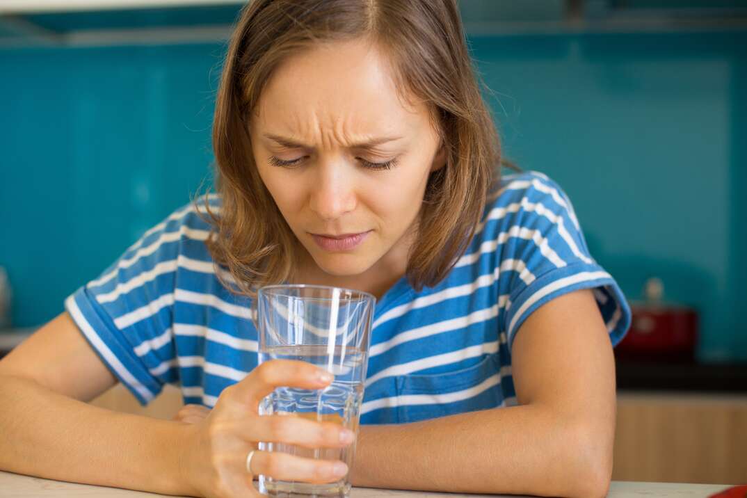 Closeup portrait of dissatisfied young beautiful woman looking into glass of water and sitting at table in kitchen. Bad quality drinking water concept. Front view.