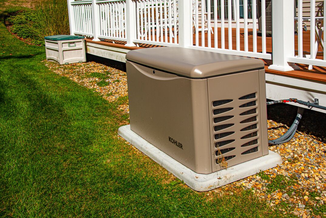 A residential backup generator sits on a concrete pad atop a green grass lawn next to the white spindles of the guard rail of the back porch of a house, generator, backup generator, residential generator, residential backup generator, electrical, electricity, power, backup power, grass, green grass, lawn, yard, backyard, concrete, concrete pad, back porch, porch
