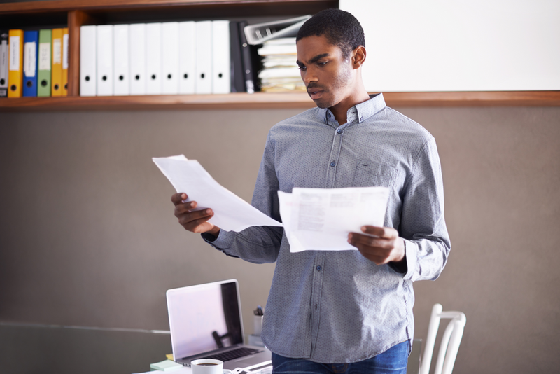 A younger man stands in an office looking at a pair of documents and ostensibly struggling to read them or make sense of what they say, contract, contracts, legal, legal contract, legal documents, documents, forms, legal forms, papers, legal papers, man, younger man, reading documents, office