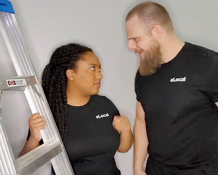 A woman in a black eLocal T-shirt stands next to a ladder as she and a man also wearing black eLocal T-shirts look at each other as if in an argument., lauren leazenby, lauren, matt schmitz, matt, leazenby, schmitz, ladder, gray wall, wall, gray, black eLocal T-shirt, elocal T-shirt, T-shirt, man, woman, argument, arguing, dispute, disputing