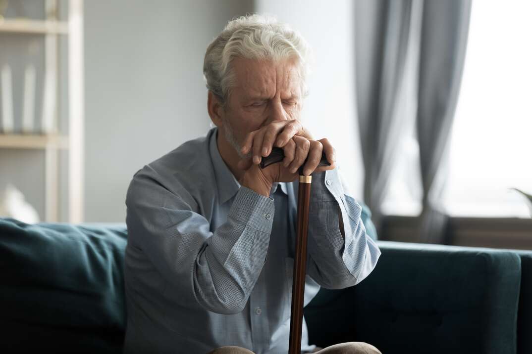 Depressed disabled retired elderly 50s grandfather hold in hands walking stick sit alone on couch looking lonely and unhappy, health problems, physically handicapped person, movement disorder concept