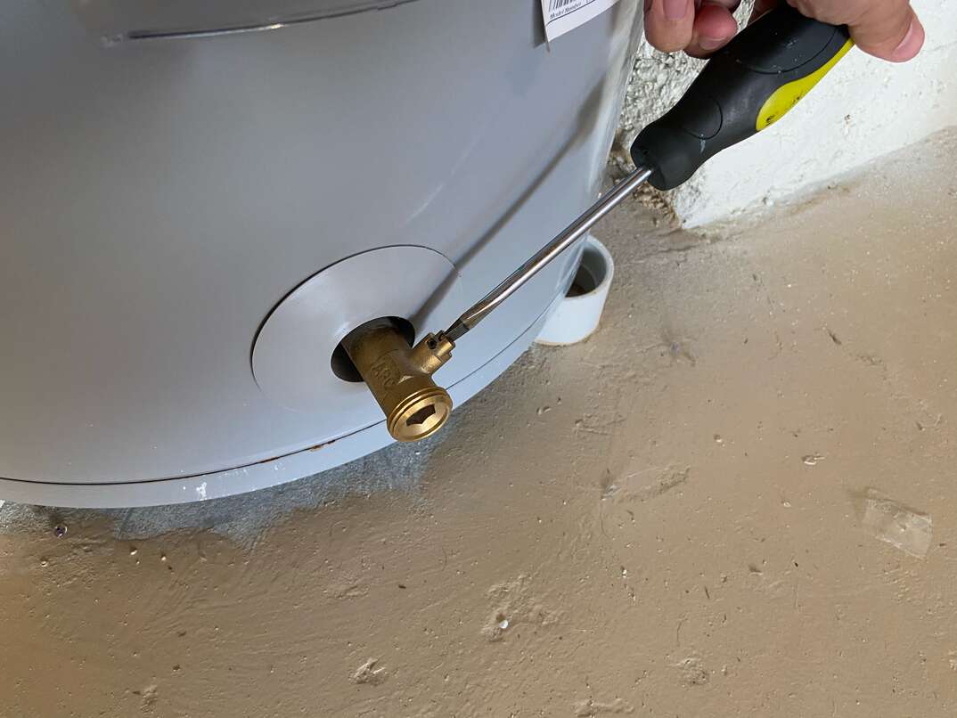 A homeowner uses a screwdriver to open the drain valve on a residential water heater 