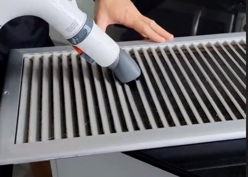 A closeup of a vacuum cleaner attachment is shown cleaning an HVAC vent with a human hand visible in the background, hvac, hvac vent, AC vent, vent, cleaning, dusting, dust, vacuum cleaner attachment, attachment, hand, human hand