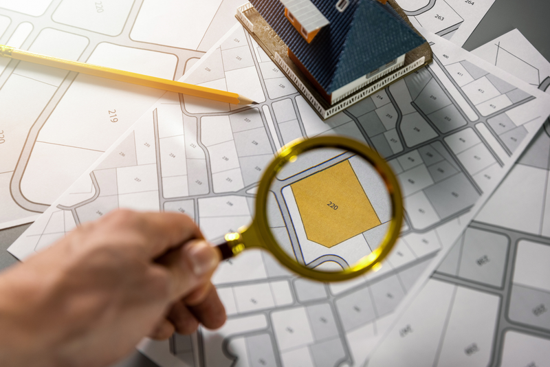 A human hand holds a magnifying glass over a residential subdivision blueprint showing property lines and property numbers and on top of that map sits a small model house and a pencil, human hand, hand, human, magnifying glass, pencil, model house, little house, small house, house, home, property lines, property blueprint, subdivision, residential neighborhood, neighborhood, map