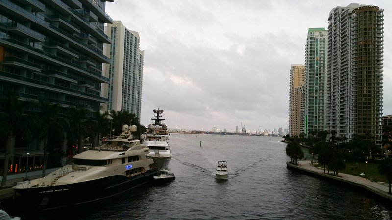 Large yachts are seen docked along the bank of a canal in Miami as a much smaller boat moves down the middle of the canal with skyscrapers lining both sides of the waterfront, boats, boat, yacht, yachts, motorboats, motorboat, motor boat, Miami, river, canal, waterway, skyline, skyscrapers, tall buildings, buildings, river banks, waterfront