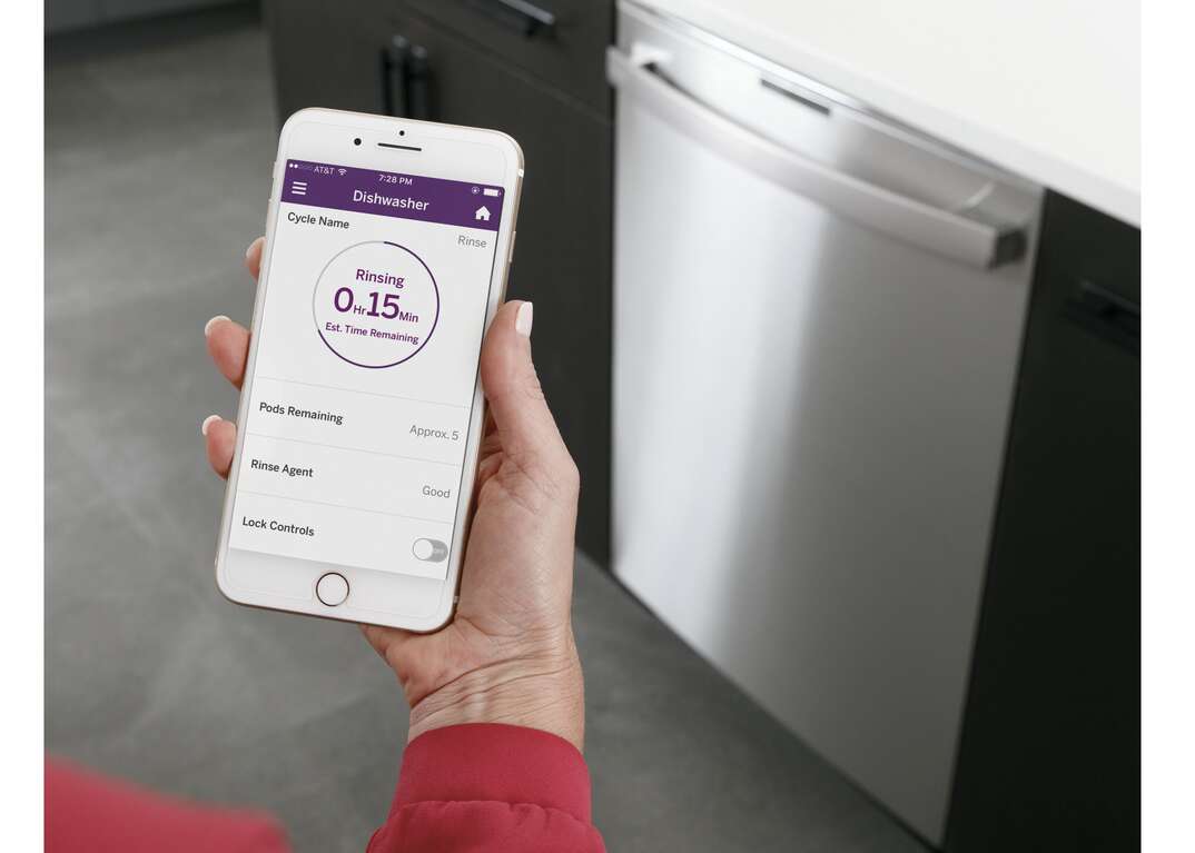 The hand of a person wearing a red shirt holds a smartphone wirelessly connected to a GE brand smart dishwasher in a kitchen