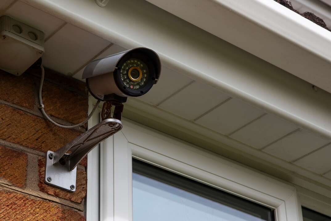 A security surveillance camera mounted on the red brick exterior wall of a house just under the white overhang of the roof and just next to a white trimmed window faces away from the home, security camera, camera, video surveillance, surveillance camera, surveillance, security, safety, red brick exterior wall, exterior wall, wall, red brick, brick, brick wall, window, roof