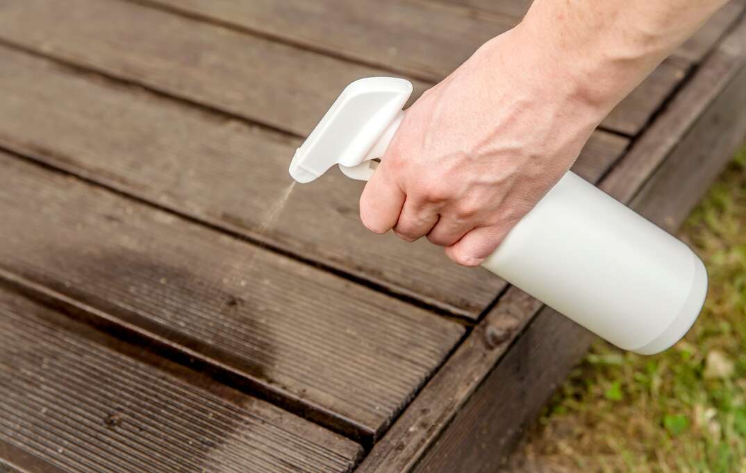 A human hand is shown in closeup using a white spray bottle to spray a liquid substance onto an outdoor brown wood plank patio with grass visible in the corner of the frame, spray bottle, bottle, spray, white spray bottle, deck, patio, wood, planks, wood plank, human hand, spraying, grass, yard, backyard