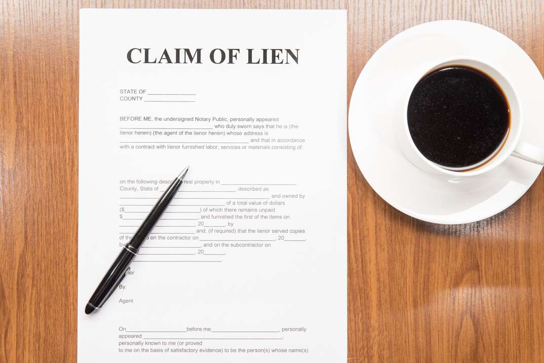 A legal document titled Claim of Lien sits on a brown woodgrain desktop with an ink pen resting on it and a white cup filled with black coffee sits in a white saucer, lien, tax lien, claim of lien, property lien, legal document, legal, document, pen, ink pen, desktop, desk, woodgrain finish, woodgrain, cup of coffee, black coffee, coffee, coffee cup, saucer