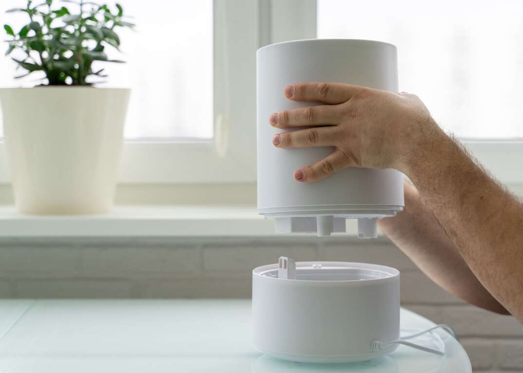 A pair of human hands is seen lifting the top off of a white tabletop humidifier for the purpose of cleaning it