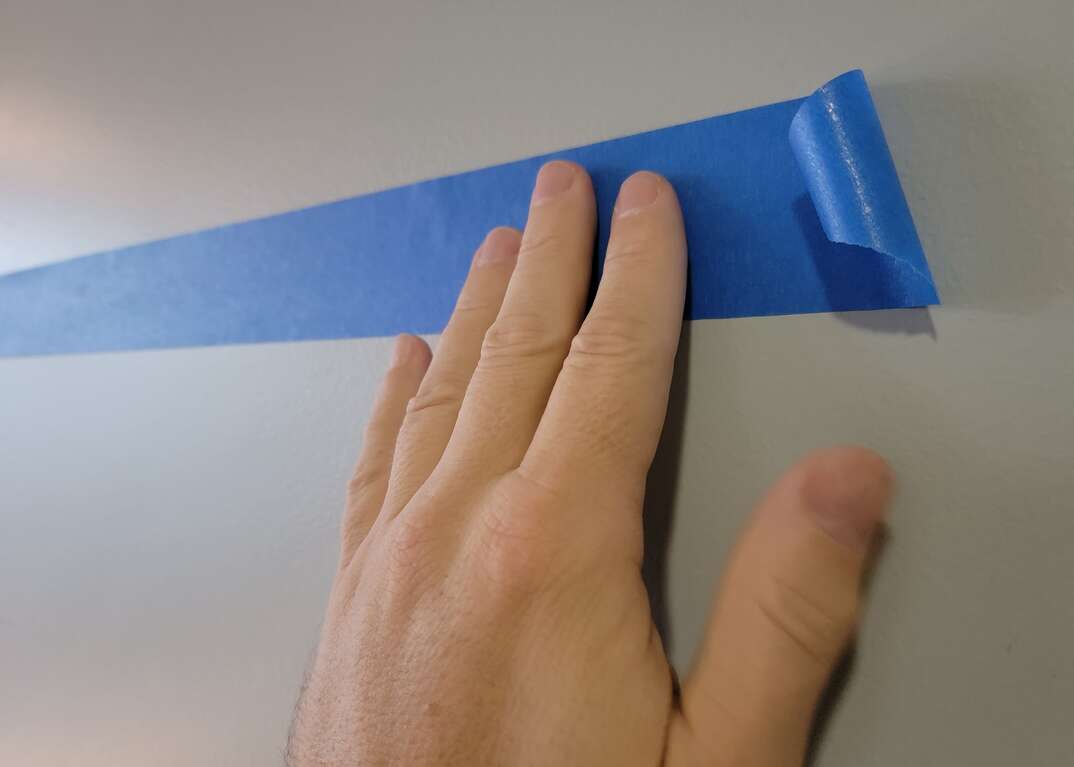 Human hand applies blue painters tape to a gray interior wall