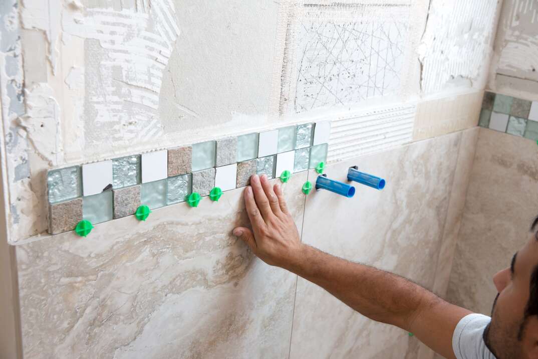 mosaic tile border made up of small square glass tiles is being adhered to a shower wall  Larger tiles are in place underneath it  The bathroom in a home The arm and head of tile worker are visible 