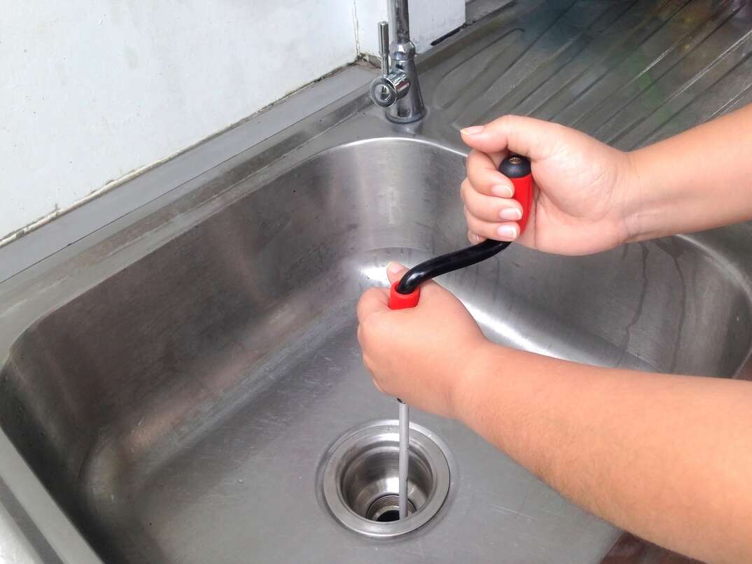 man rotating a drain snake into a kitchen sink to unclog the pipe