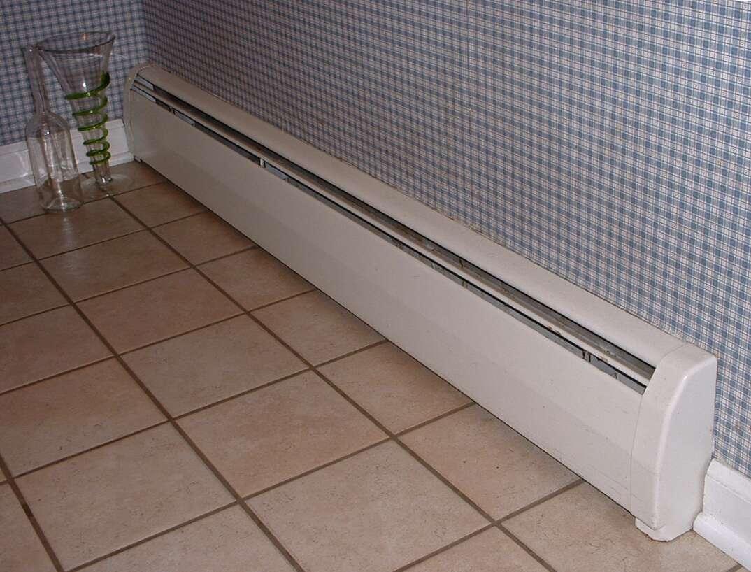 A white colored baseboard heater is shown affixed to the lower portion of the wall in a room with a tile floor, tile floor, tile, baseboard heater, baseboard, heater, radiator, HVAC, heat