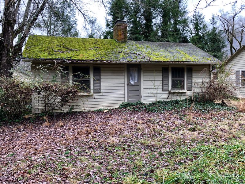An old one story house in disrepair sits abandoned in a wooded area with green mossy vegetation growing on the roof and dead leaves on the ground and poorly maintained shrubbery in front