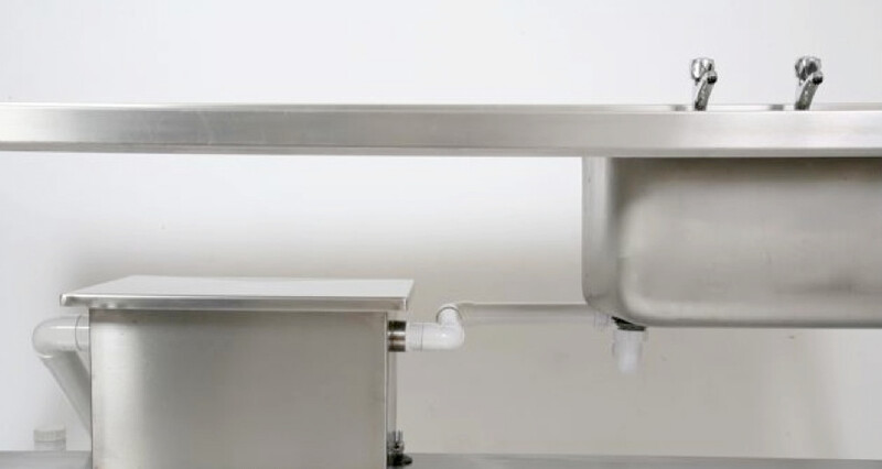 A grease trap is shown positioned underneath a kitchen countertop with a sink that is connected to the trap via PVC pipe in an all white room, grease trap, trap, grease, sink, kitchen sink, kitchen counter, kitchen countertop, countertop, counter, white, all white, white background, grease trap underneath sink, faucet