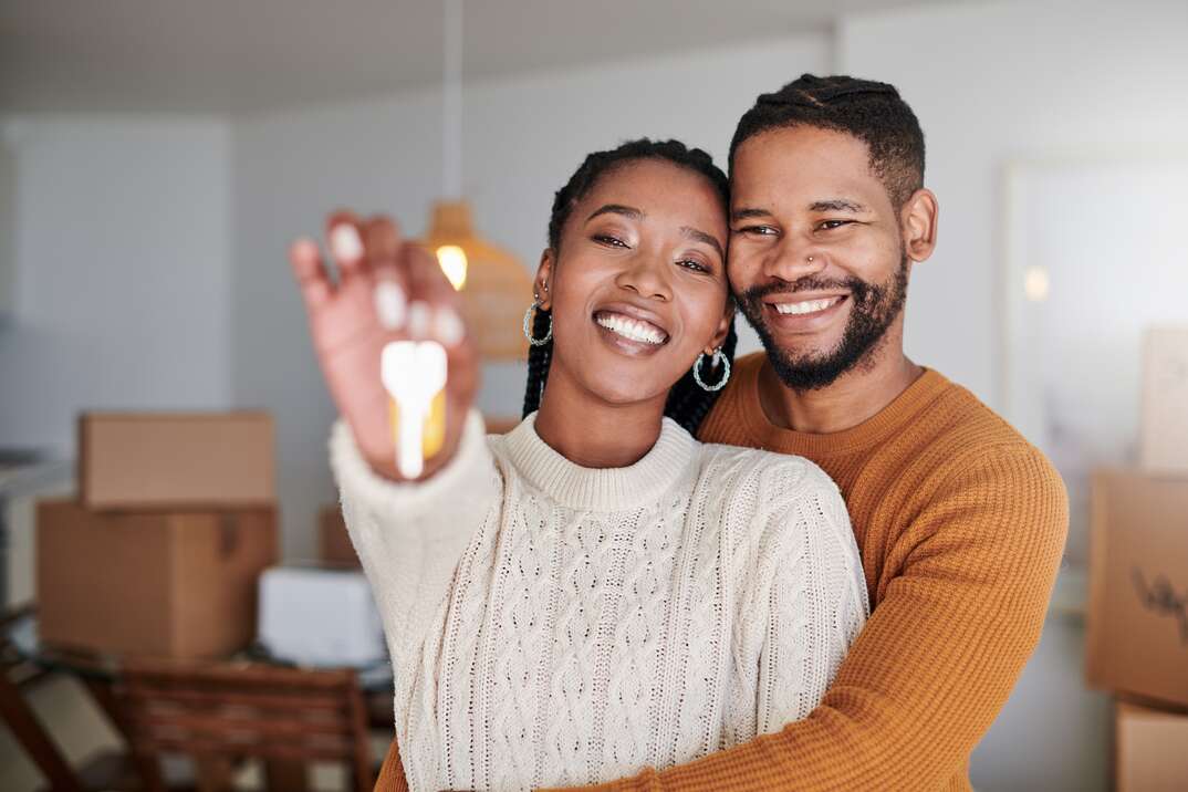 A male and female couple embrace as the woman holds out a key in front of her with boxes in the background waiting to be unpacked in their new residence, male, female, male and female, male and female couple, key, house keys, new home, home, house, boxes, unpacking, moving in