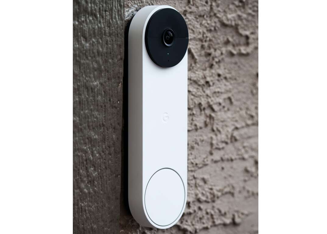 A white Google Nest Doorbell video security device with a black circular camera is affixed to a brown stucco exterior wall.