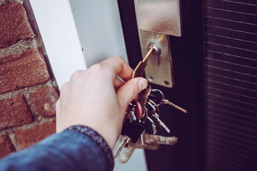 A closeup of a male hand unlocks the front door deadbolt of a red brick residence using a key attached to a keyring with many keys attached to it, male hand, keyring, key ring, keychain, key chain, keys, house keys, unlocking door, unlocking, door, front door, deadbolt, red brick residence, red brick, brick, residence, apartment, home, house, front door, door