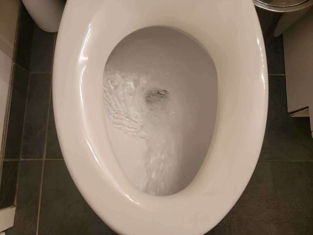 A birds eye view of a white toilet with the seat down as it flushes, flushing toilet, toilet, flush, flushing, bathroom, restroom, residential toilet, toilet bowl, toilet seat, tile floor, porcelain toilet, porcelain, water