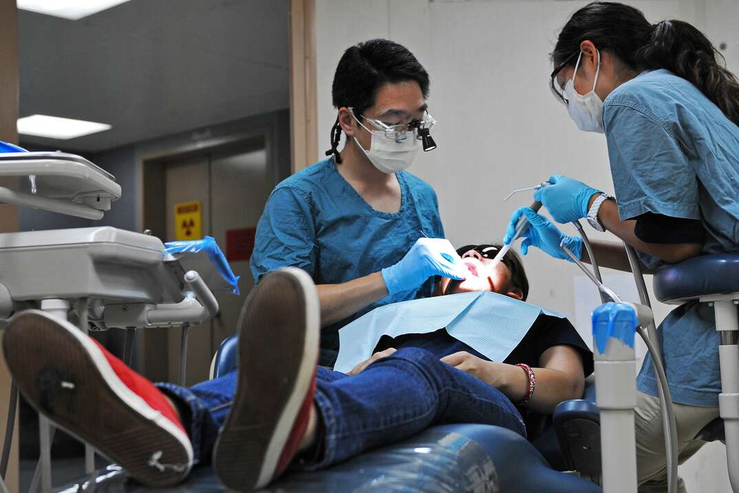 Two dentist dressed in medical scrubs and surgical masks perform a root canal on a male patient who is reclined in a dentist chair in a dentist office, dental, dentist, dentistry, dental health, medical, medical procedure, surgery
