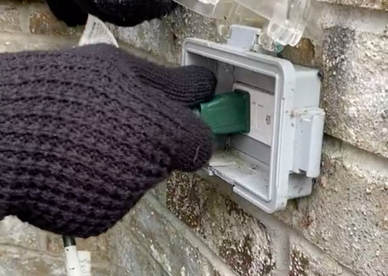 Human hand wearing a black glove plugs in a green plug for holiday lights into an exterior wall outlet housed in a plastic outlet cover, black glove, hand in black glove, glove, hand, human hand, green plug, plug, outlet, exterior outlet, home outlet, household outlet, wall outlet, outdoor outlet cover, outlet cover, holiday lights