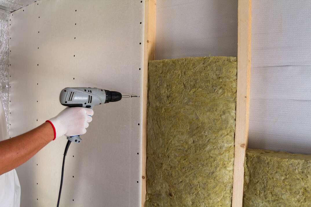 Close-up of worker hand with electric screwdriver fastening drywall to wooden timber frame with insulation mineral rock wool staff. Warm home