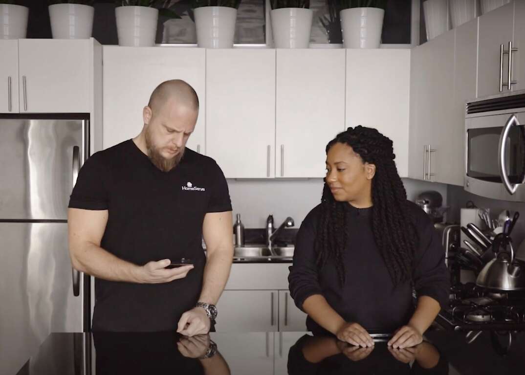 HomeServe Managing Editor Matt Schmitz and HomeServe Editor Lauren Leazenby stand at a black granite countertop in a kitchen with white cabinets as Matt checks his smartphone and Lauren looks on, Matt Schmitz, Lauren Leazenby, male, female, HomeServe employees, black granite countertop, kitchen, kitchen cabinets, stainless steel refrigerator, refrigerator, fridge, appliances, kitchen sink, microwave, stove, teapot, tea kettle, potted plants, smartphone, cell phone, Samsung smartphone, Android device