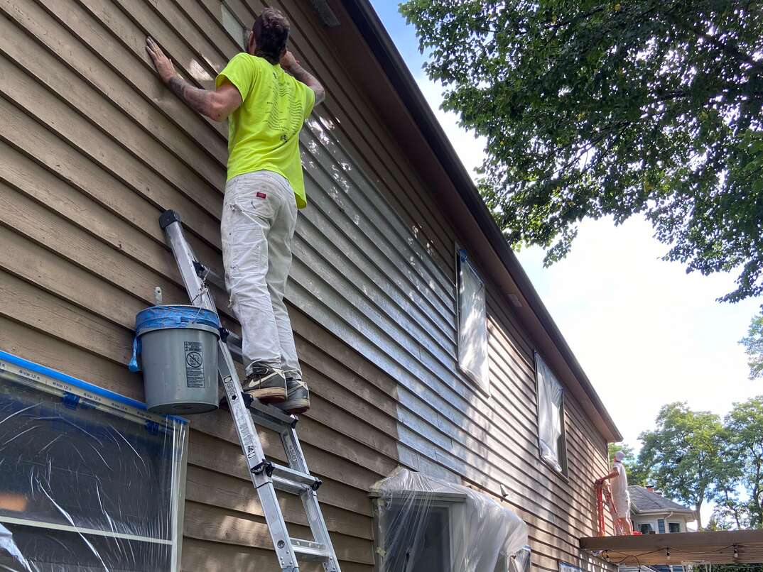 man in a yellow shirt standing tall on a ladder painting a house