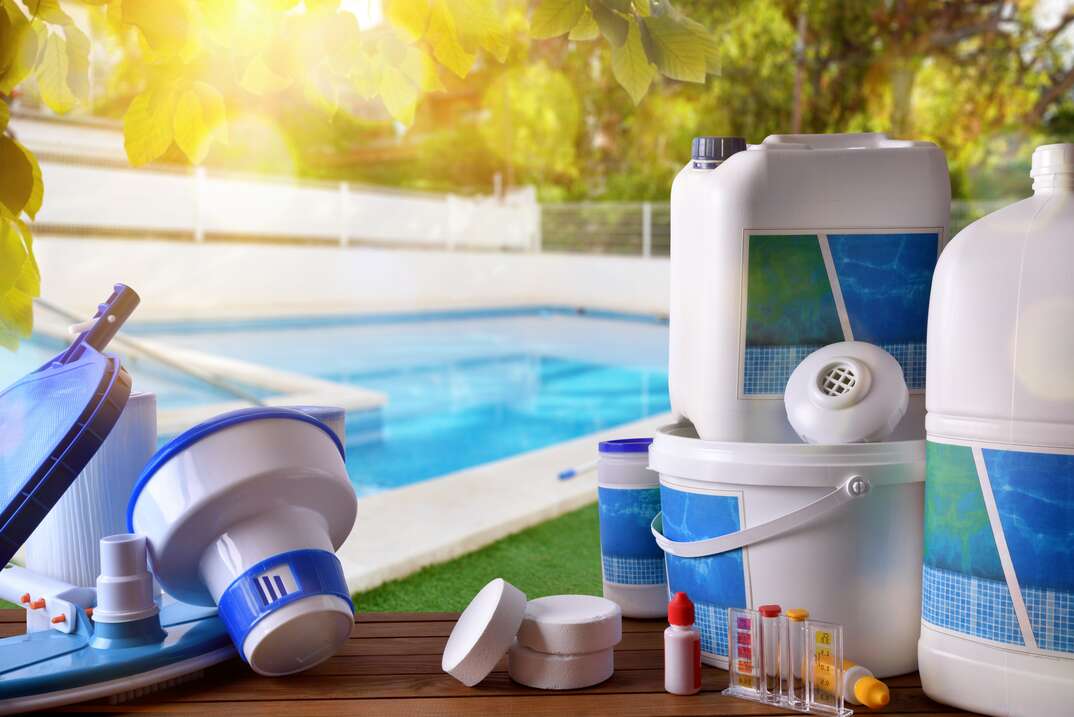 A full complement of swimming pool chemicals and related implements sits in the foreground with the blue water of an in ground swimming pool visible in the background as well as greenery, swimming pool chemicals, swimming pool, pool, pool chemicals, chemicals, cleaning chemicals, chlorine, pH balance, blue water, water, trees, greenery, shrubs, swimming, chemical testing kit, pool testing kit