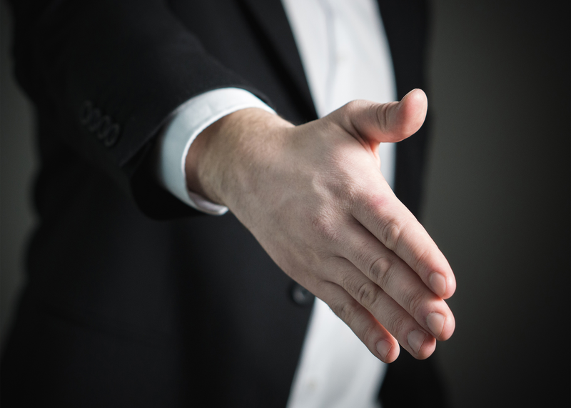 A closeup photo shows a man wearing a business suit extending his hand into the foreground to offer it for a handshake ostensibly for the purpose of closing a legal or business negotiation, negotiation, lawyer, law, attorney, legal, handshake, shaking hands, business suit, suit, attorney, business, businessman