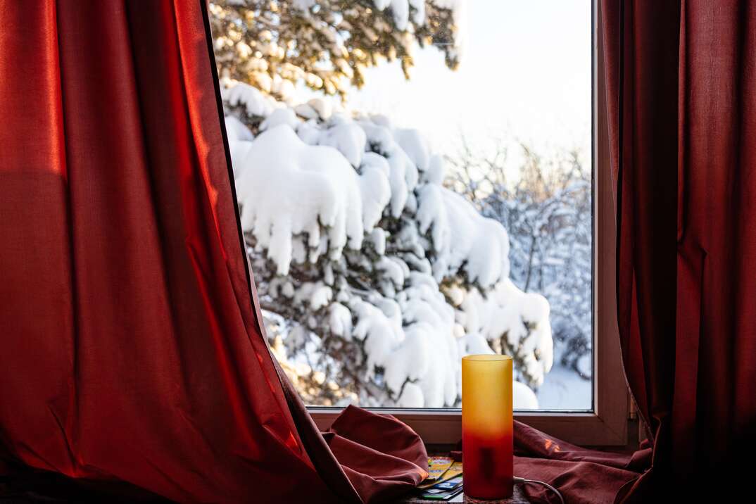 A view from inside a house through a window with heavy red curtains and a candle in front of it looks outside to a snow covered tree