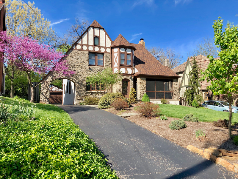 A black asphalt driveway leads up to a large house surrounded by lush greenery and landscaping set against a blue sky, asphalt, asphalt driveway, blacktop, paved, pavement, house, big house, house, nice house, landscaping, greenery, front yard, yard, lawn, front lawn