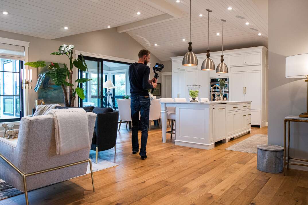A man dressed in dark clothing uses a camera on a gimbal to photograph the interior of a very tidy and well designed and well decorated house with new looking hardwood floors and a large island countertop, staged house, selling house, house selling, real estate, taking pictures, photos, photographer, man, male photographer, living room, kitchen, hardwood floors, hardwood, island countertop, island, countertop, hanging light fixtures, hanging lights, lights, light fixtures, light, fixtures, beautiful home