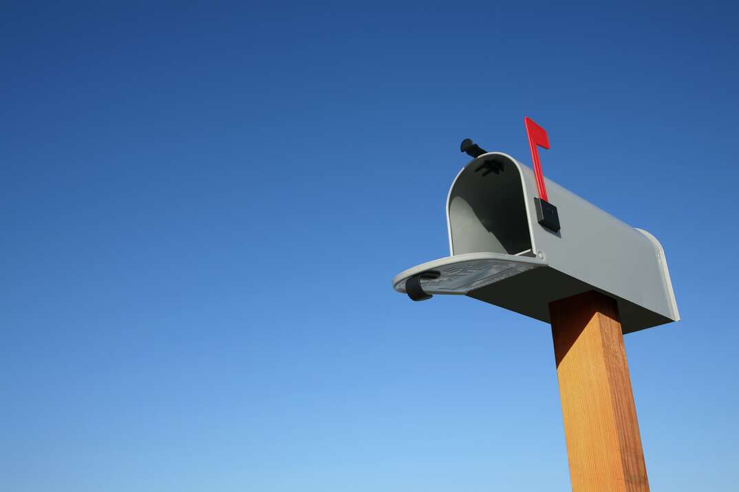A gray mailbox sits with its red flag up and door open atop a brown wooden post against the backdrop of a cloudless blue sky