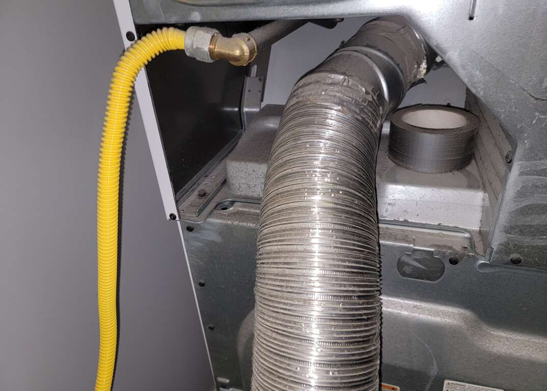 Rear view of a dryer with the dryer vent tube and a yellow hose extending from the appliance to the bottom of the photo, and a roll of duct tape sitting on a shelf next to the dryer vent tube.