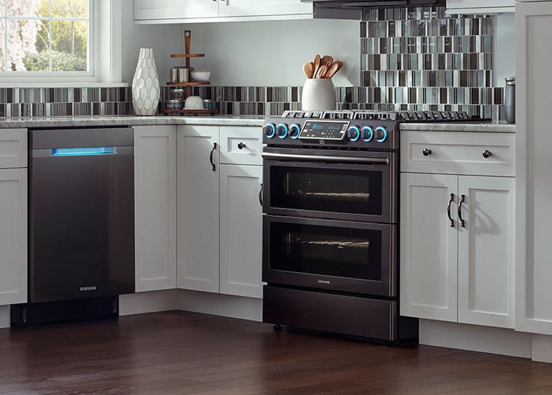 Modern kitchen featuring a Samsung smart stainless steel range with glowing blue knobs a brown wood floor white cabinetry a stainless steel dishwasher a tile backsplash and a window with natural light coming in