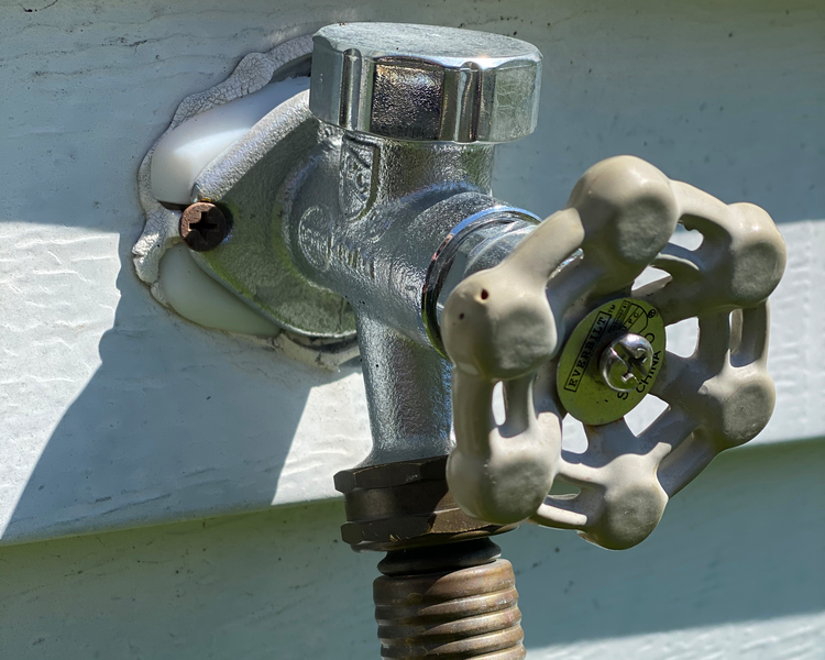 A closeup of an outdoor faucet also known as a hose Bibb is shown with the hexagonal shaped handle and chrome Bibb extending from white house siding, white siding, siding, hexagon, hexagonal, faucet, outdoor faucet, outdoor, outside, chrome, metallic, hose bibb, bibb, hose