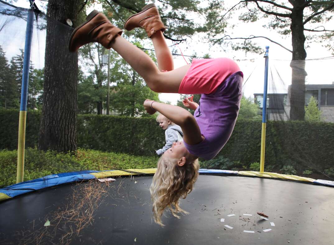 A young girl hangs upside down in mid air as she bounces on a large round trampoline in a residential yard with another young child in the background, yard, back yard, backyard, green grass, lawn, trees, tree, residential, upside-down, mid-air, jumping on trampoline, jumping, trampoline, bouncing, bouncing on trampoline, fun, having fun, recreation, recreational