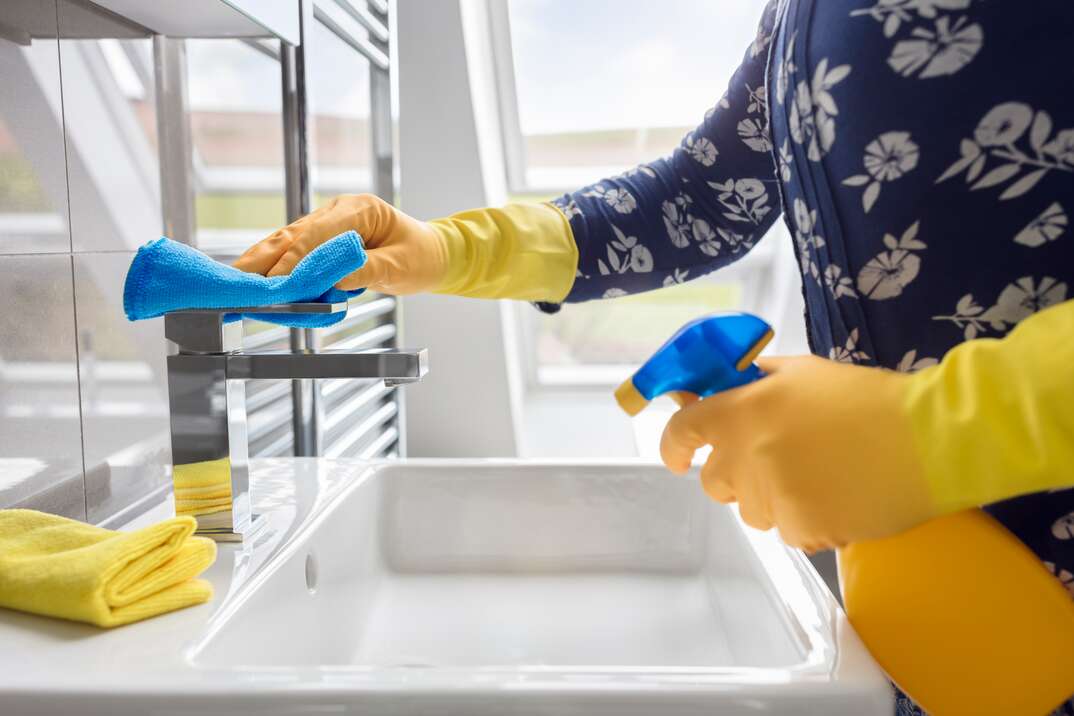woman wearing yellow rubber gloves sprays cleaner on to a bathroom faucet