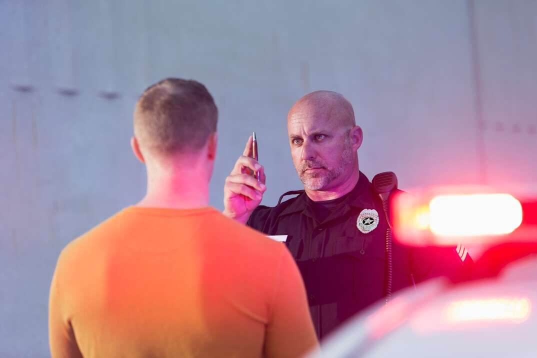 A police officer administers a field sobriety test to a stopped driver standing in front of him with his back to the viewer as the red patrol car lights flash in the foreground, field sobriety test, driver, drunk driver, drunk, DUI, driving under the influence, DWI, driving while intoxicated, police officer, police, officer, patrol car, car, police car, police lights, flashing red lights, lights, flashing, drunk, intoxicated, drinking and driving