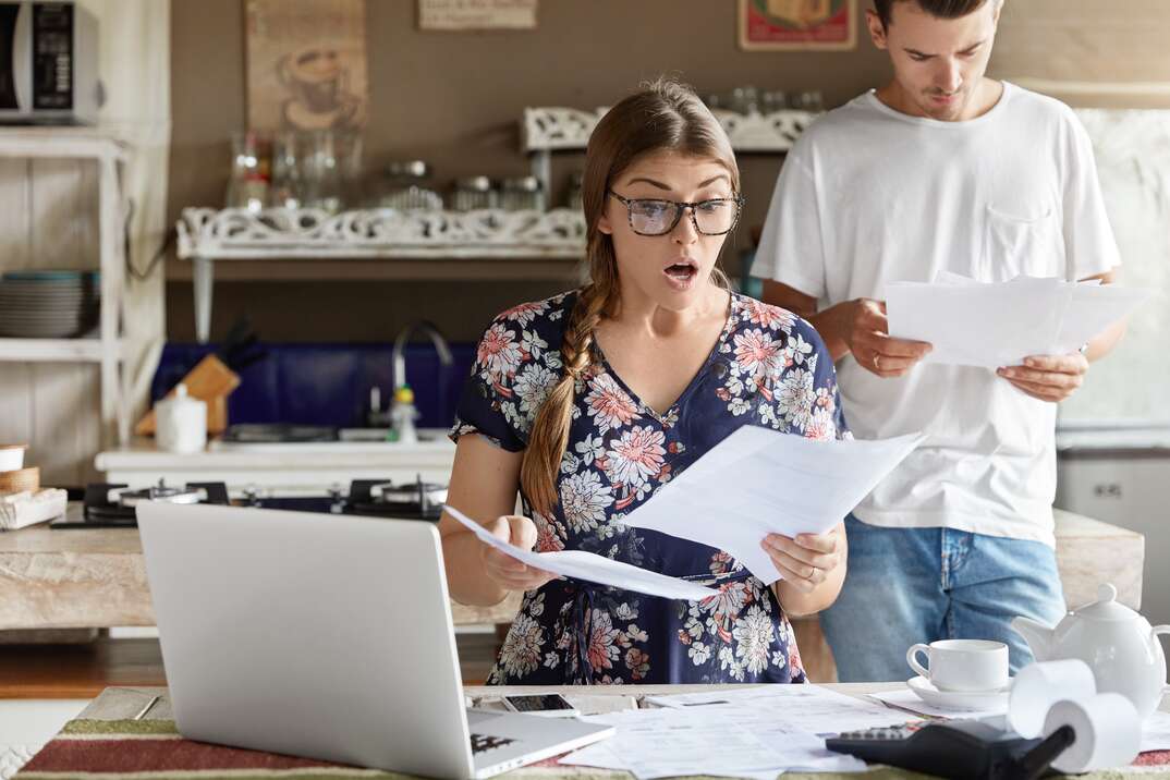 Young female looks with shocked expression into papers, recieve high taxes and had to pay much money for bills, sits against kitchen interior, her husband stands behinds, studies documents attentively